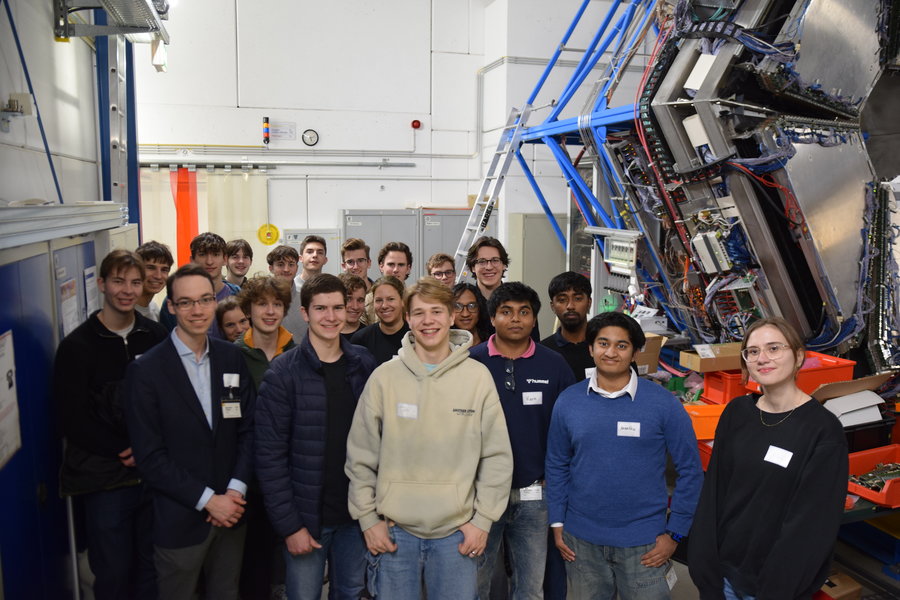 Die Teilnehmenden der ALICE-Masterclass 2026 Eine Gruppe von Personen steht in einem großen Raum. Am rechten Rand ist ein haushoher technischer Aufbau zu sehen.