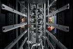 KI-System beim Einbau im Green IT Cube Perspektivisch durch ein schwarzes Computerrack geschaut, sind zwei Menschen sichtbar, die an Rohrleitungen arbeiten.