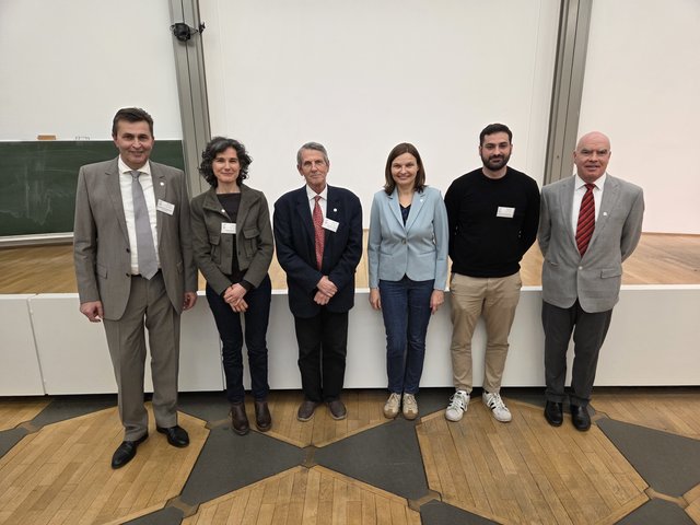 Vier Männer und eine Frau stehen vor einer Bühne. Vier Männer und eine Frau stehen vor einer Bühne.