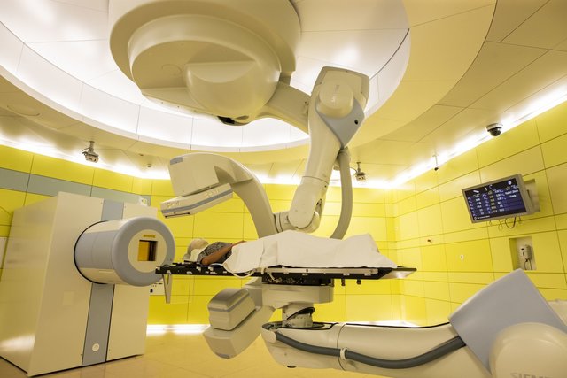 Eine Person liegt auf einer von einem Roboter gehaltenen Patientenliege vor einem zylindrischen Strahlaustrittsfenster. Eine Person liegt auf einer von einem Roboter gehaltenen Patientenliege vor einem zylindrischen Strahlaustrittsfenster.