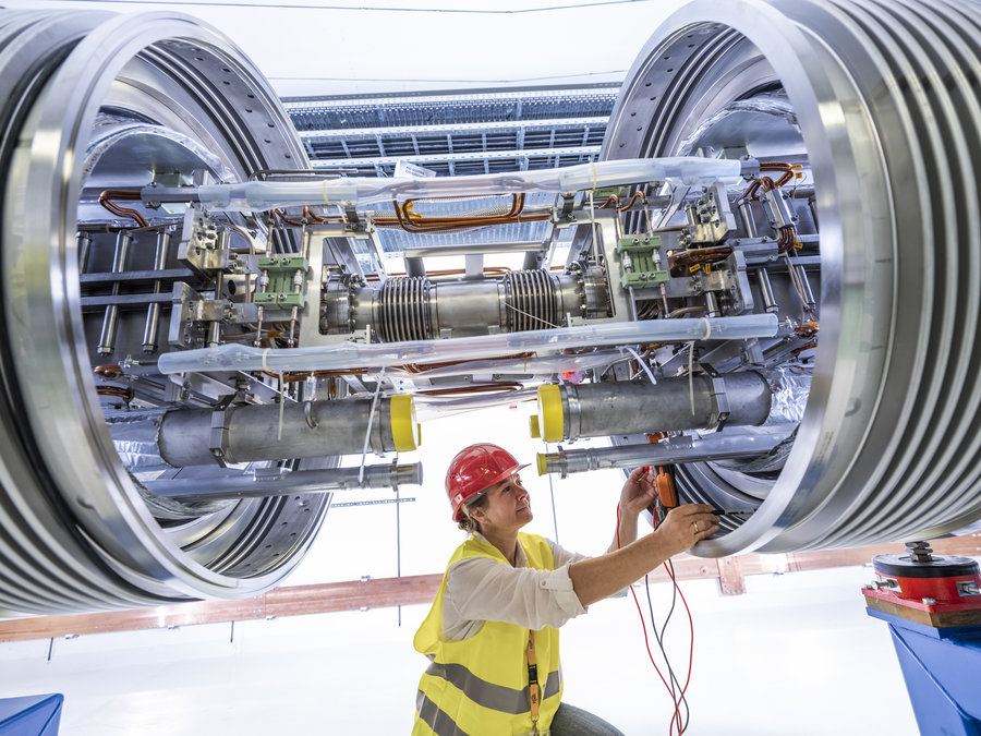 Blick in die Zukunft – Magnetinstallation am Ringbeschleuniger SIS100 von FAIR Eine Frau mit gelber Schutzweste und rotem Helm kniet unterhalb von zwei großen Röhren und verbindet die Kabel und Leitungen zwischen beiden.