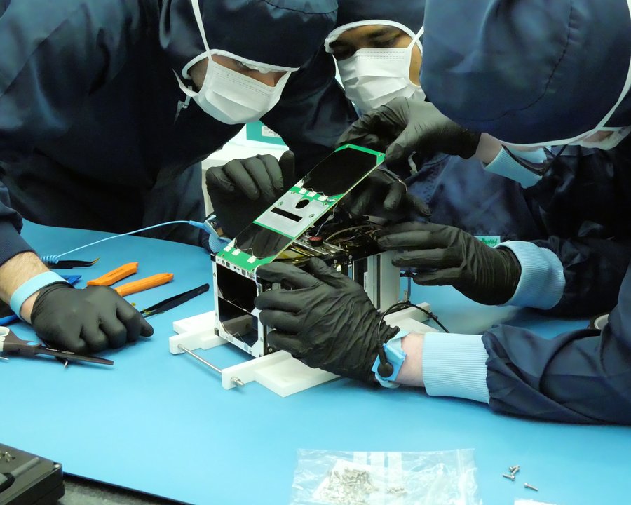 Der TRACE-Satellit wird im GSI/FAIR-Detektorlabor zusammengebaut Mehrere Personen in Schutzanzügen beugen sich über einen rechteckigen Kasten mit Elektronik.